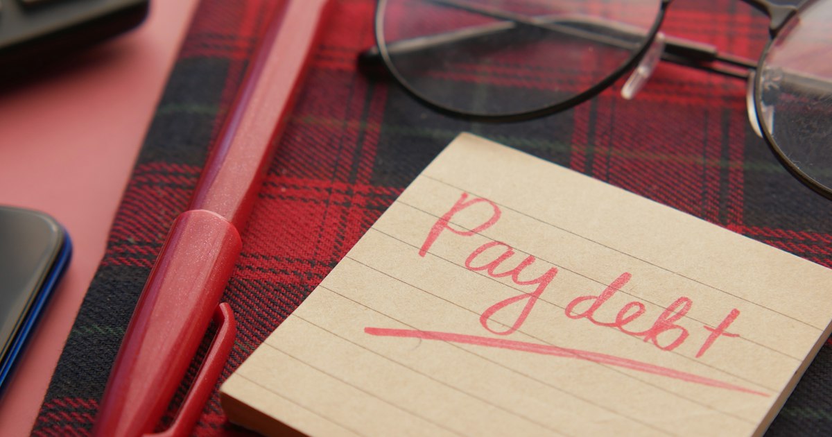 Записка «Pay debt» на столе рядом с очками и ручкой — напоминание погасить долг